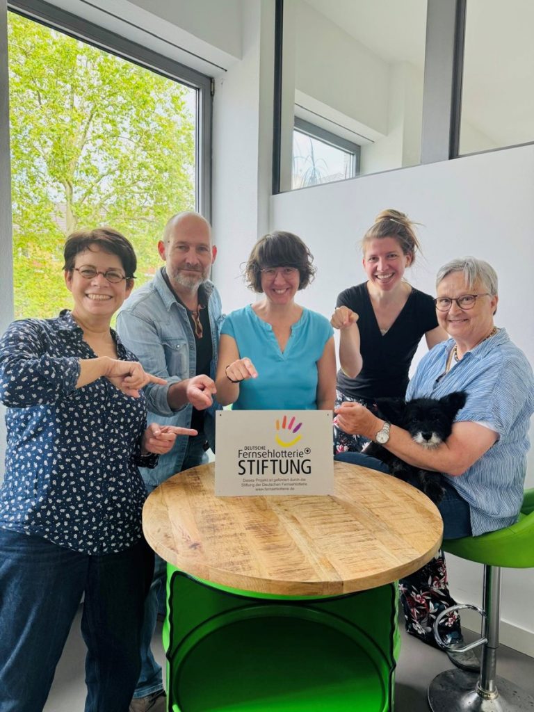 Das Team des Buddy-Projekts und Karin Ohler von der CCK zeigen auf ein Schild der Deutschen Fernsehlotterie 